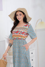 Femme souriante en robe à motifs floraux colorés, portant un chapeau de paille, posant dans une pièce lumineuse. Mode estivale, élégance décontractée.