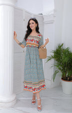 Femme souriante en robe longue à motifs floraux, tenant un sac en osier, posant près d'une colonne blanche et d'une plante verte. Mode estivale élégante.