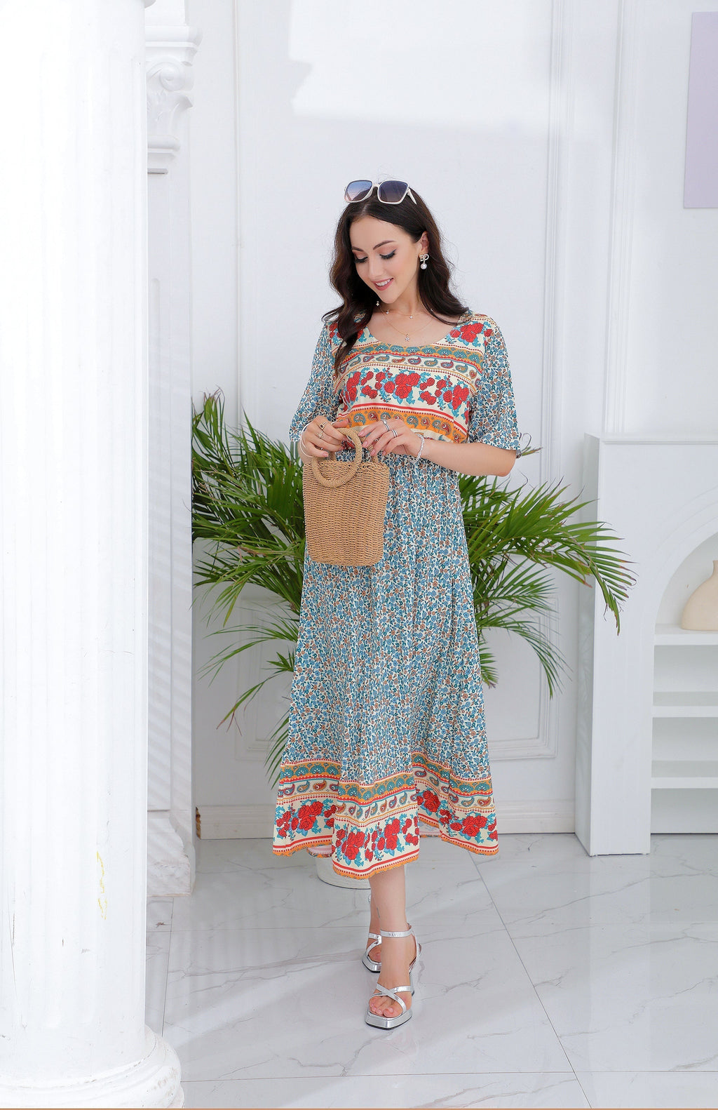 Femme en robe bohème fleurie bleue et rouge, tenant un sac en osier, posant dans un intérieur lumineux avec plantes vertes. Mode estivale élégante.