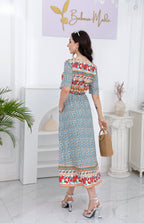 Femme en robe bohème à motifs floraux, portant un sac en osier, debout dans un intérieur moderne. Mode estivale, style bohème chic.