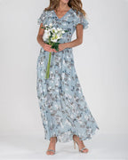 Robe longue fleurie bleue avec manches courtes, portée par une femme tenant un bouquet de fleurs blanches. Mode élégante, tenue estivale chic.