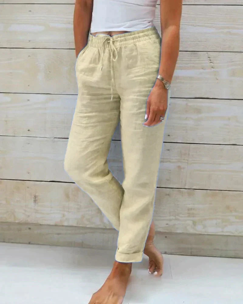 Pantalon en lin beige pour femme, taille élastique avec cordon, poches latérales, style décontracté, porté avec un haut blanc, fond en bois clair.