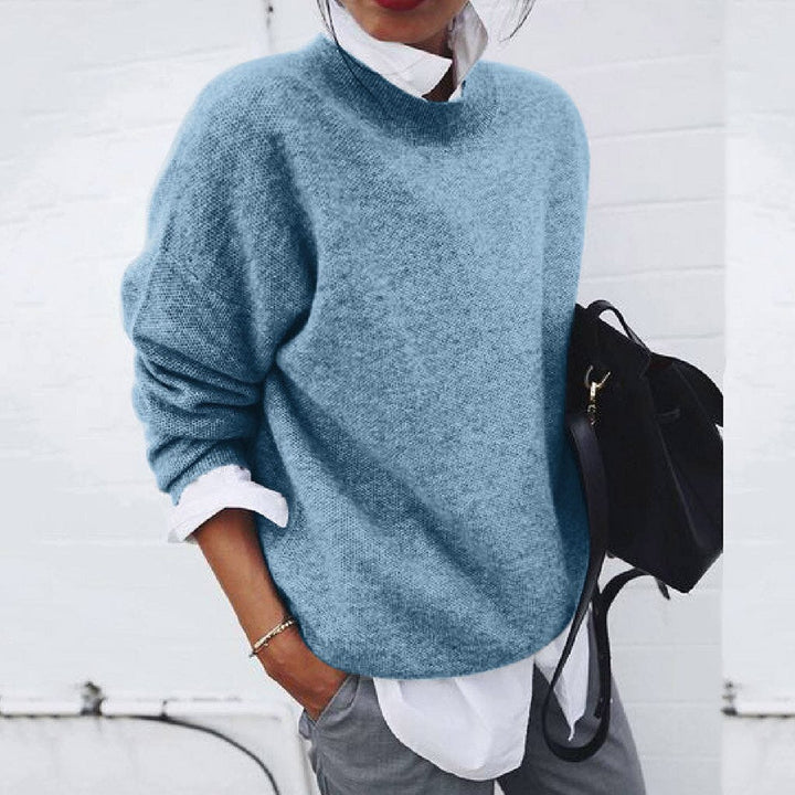 Femme portant un pull bleu ample, chemise blanche et pantalon gris, tenant un sac noir. Mode décontractée, tendance automne-hiver.