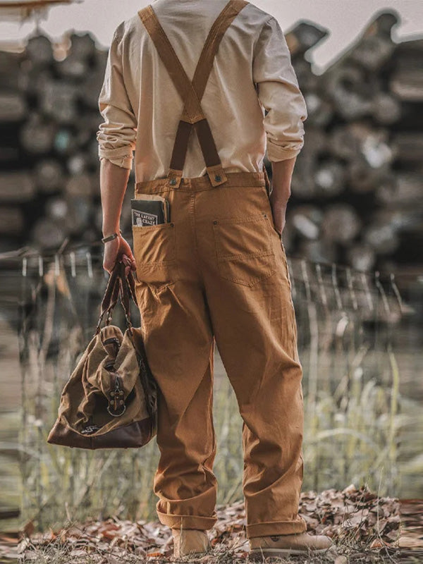 Homme de dos portant une chemise blanche et un pantalon à bretelles marron, tenant un sac en toile, devant un fond de bûches empilées.