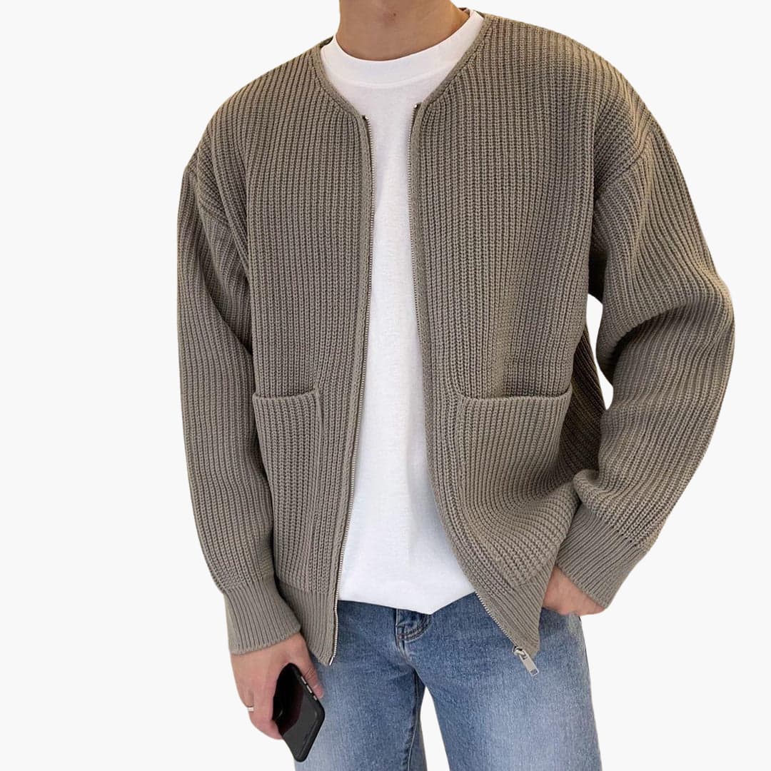 Homme portant un cardigan en tricot beige avec poches, t-shirt blanc et jean bleu, tenant un smartphone. Mode décontractée, style automne-hiver.