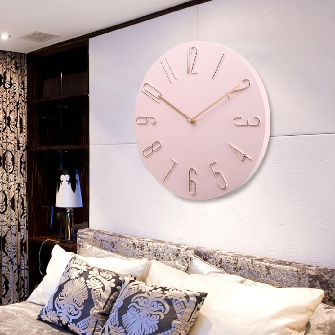 Horloge murale rose moderne avec chiffres dorés, accrochée dans une chambre élégante avec lit et coussins décoratifs, éclairage doux.