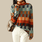 Femme portant un pull à carreaux multicolore avec col roulé, sac à bandoulière marron, et pantalon blanc. Mode automne-hiver tendance.