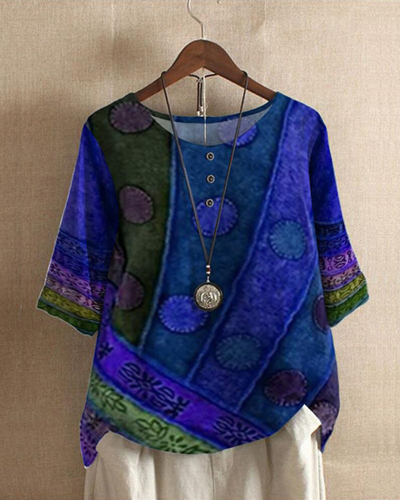 Blouse bohème bleue à motifs ethniques, manches trois-quarts, col rond avec boutons, suspendue sur un cintre, collier pendentif argenté.