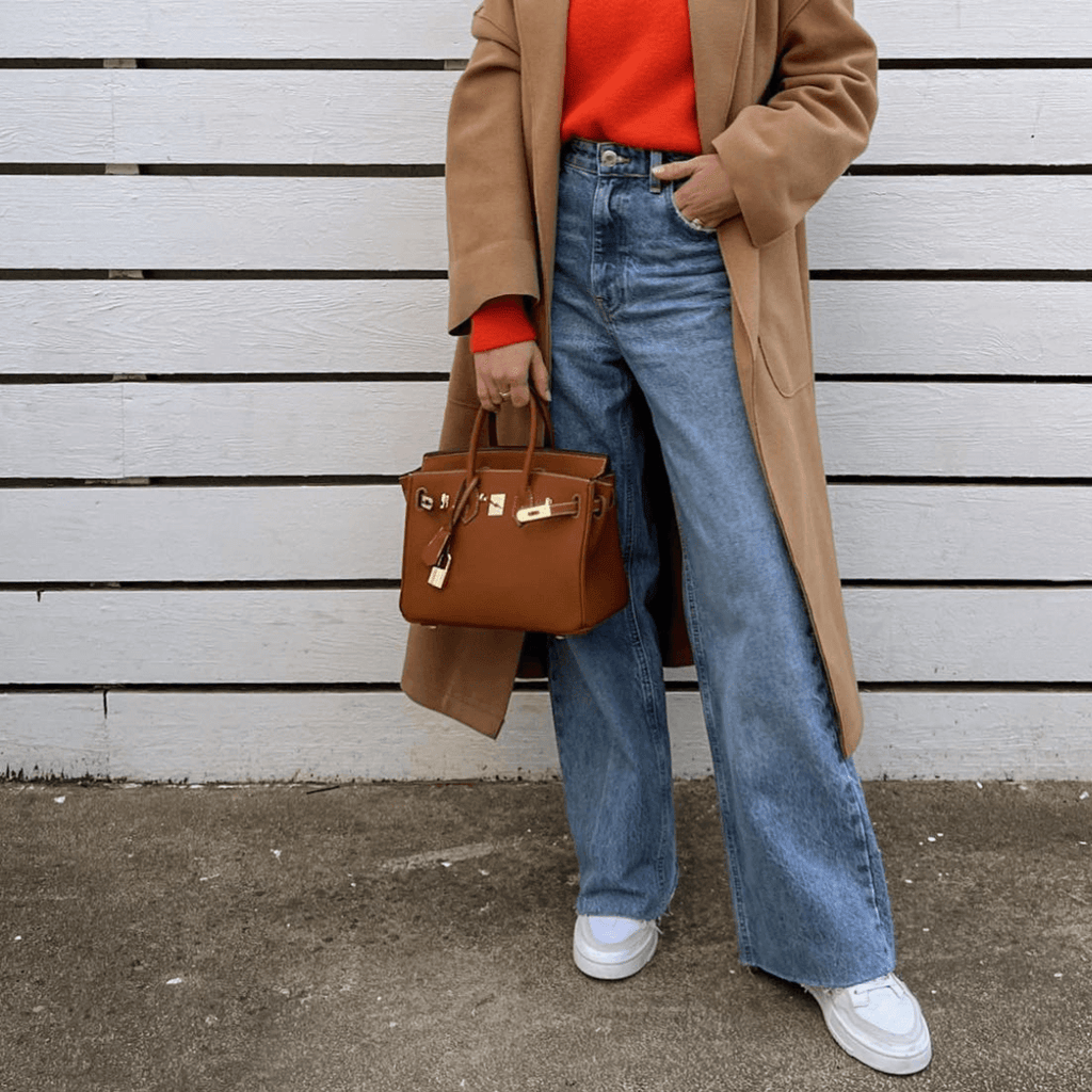 Femme en manteau camel, pull rouge, jean large et baskets blanches, tenant un sac à main marron devant une clôture blanche. Mode automne chic.