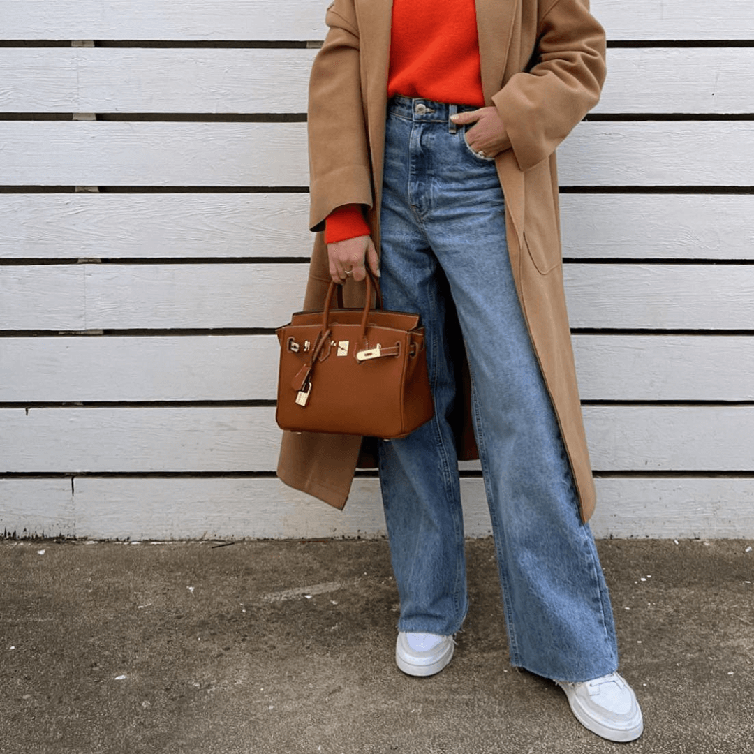 Femme en manteau camel, pull rouge, jean large et baskets blanches, tenant un sac à main marron devant une clôture blanche. Mode automne chic.