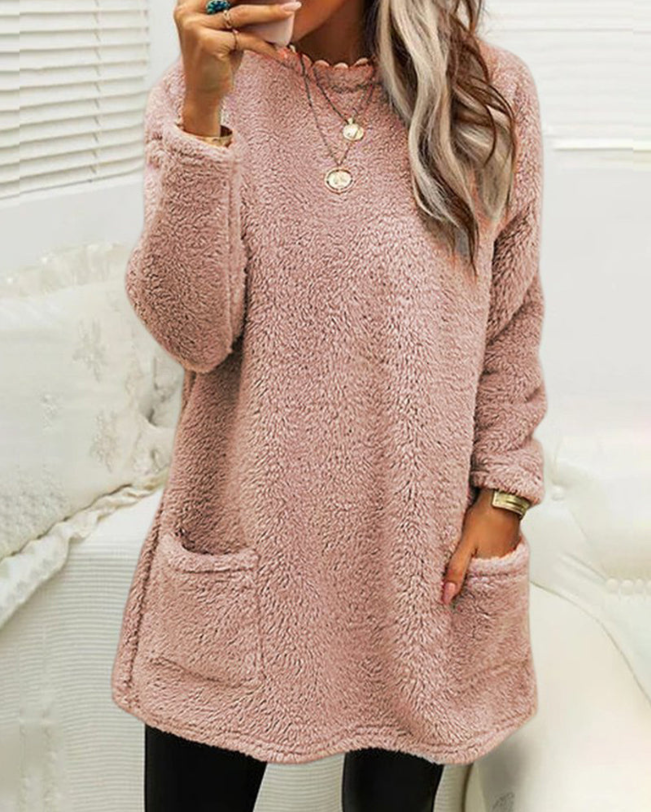 Femme portant un pull en laine rose avec poches, col rond, manches longues, posant dans un intérieur lumineux. Mode hiver, tenue confortable.