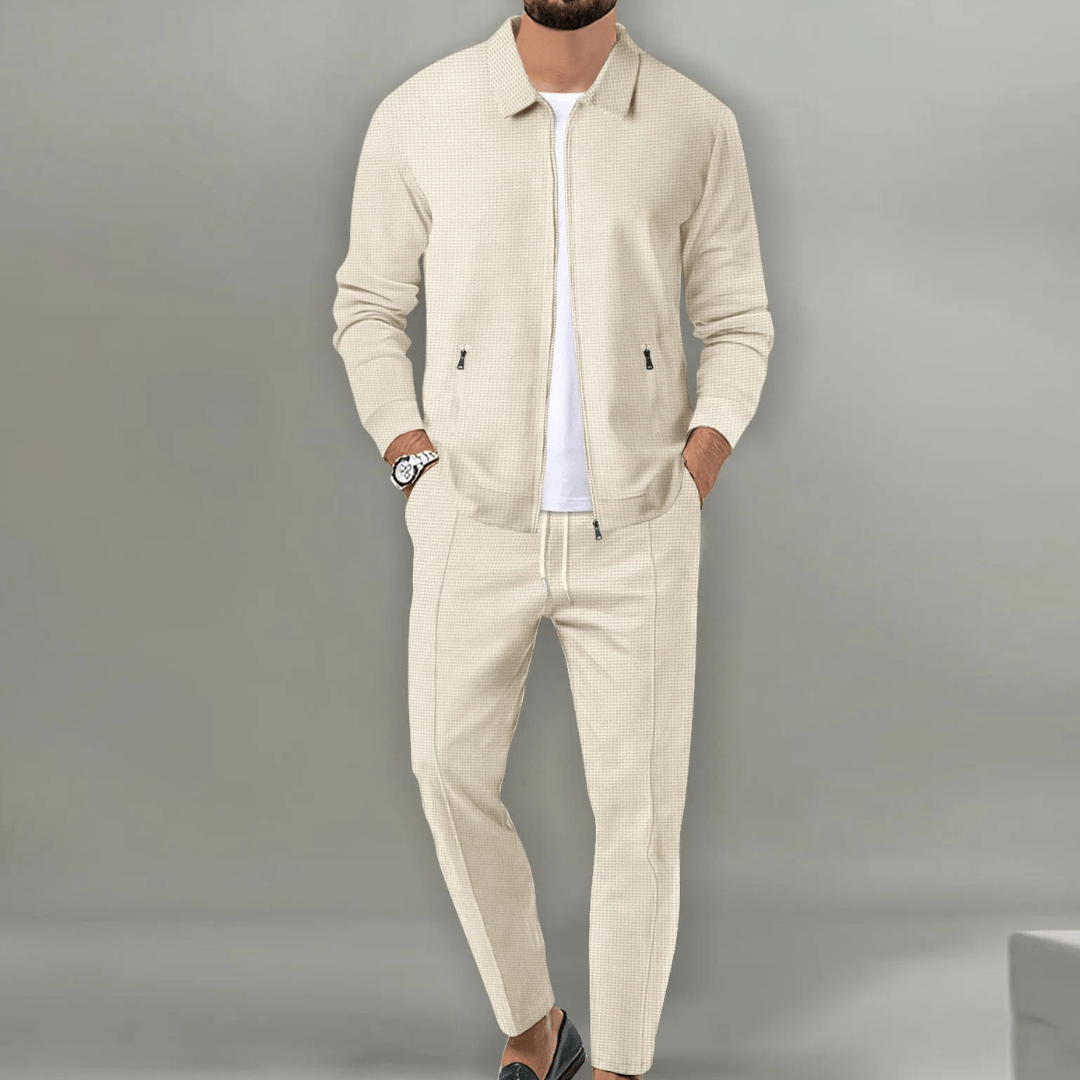 Homme portant un ensemble beige élégant, veste et pantalon assortis, sur fond gris. Mode masculine, tenue décontractée chic, style moderne.