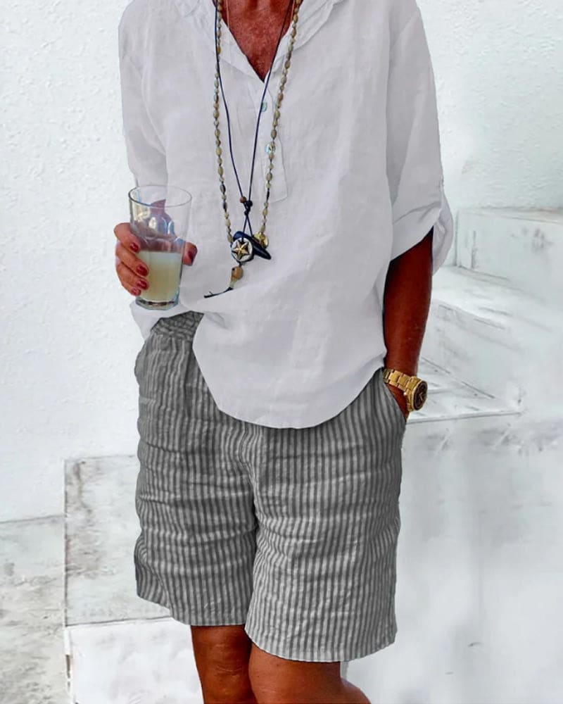 Homme en chemise blanche et short rayé gris, tenant un verre. Style décontracté estival, accessoires en bois et montre dorée. Mode masculine tendance.