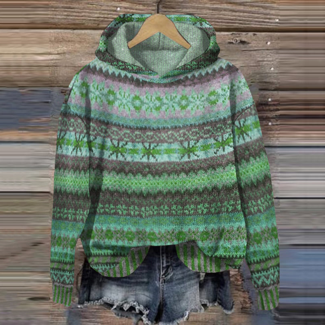 Pull à capuche vert à motifs nordiques suspendu sur un cintre en bois, devant un fond en bois rustique, accompagné d'un short en jean décontracté.