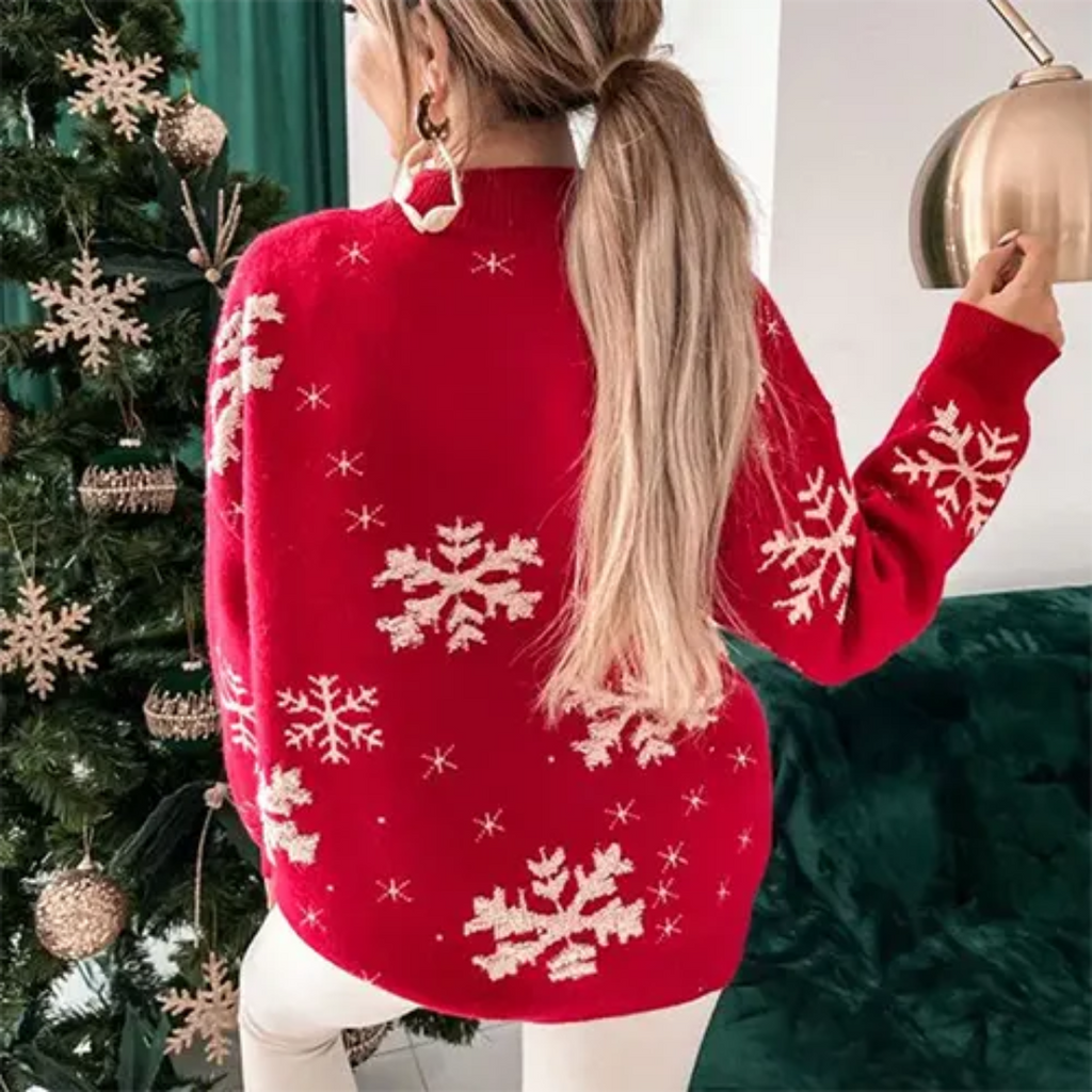 Femme portant un pull rouge avec motifs flocons de neige, debout près d'un sapin de Noël décoré. Mode hiver, tenue festive, ambiance Noël.