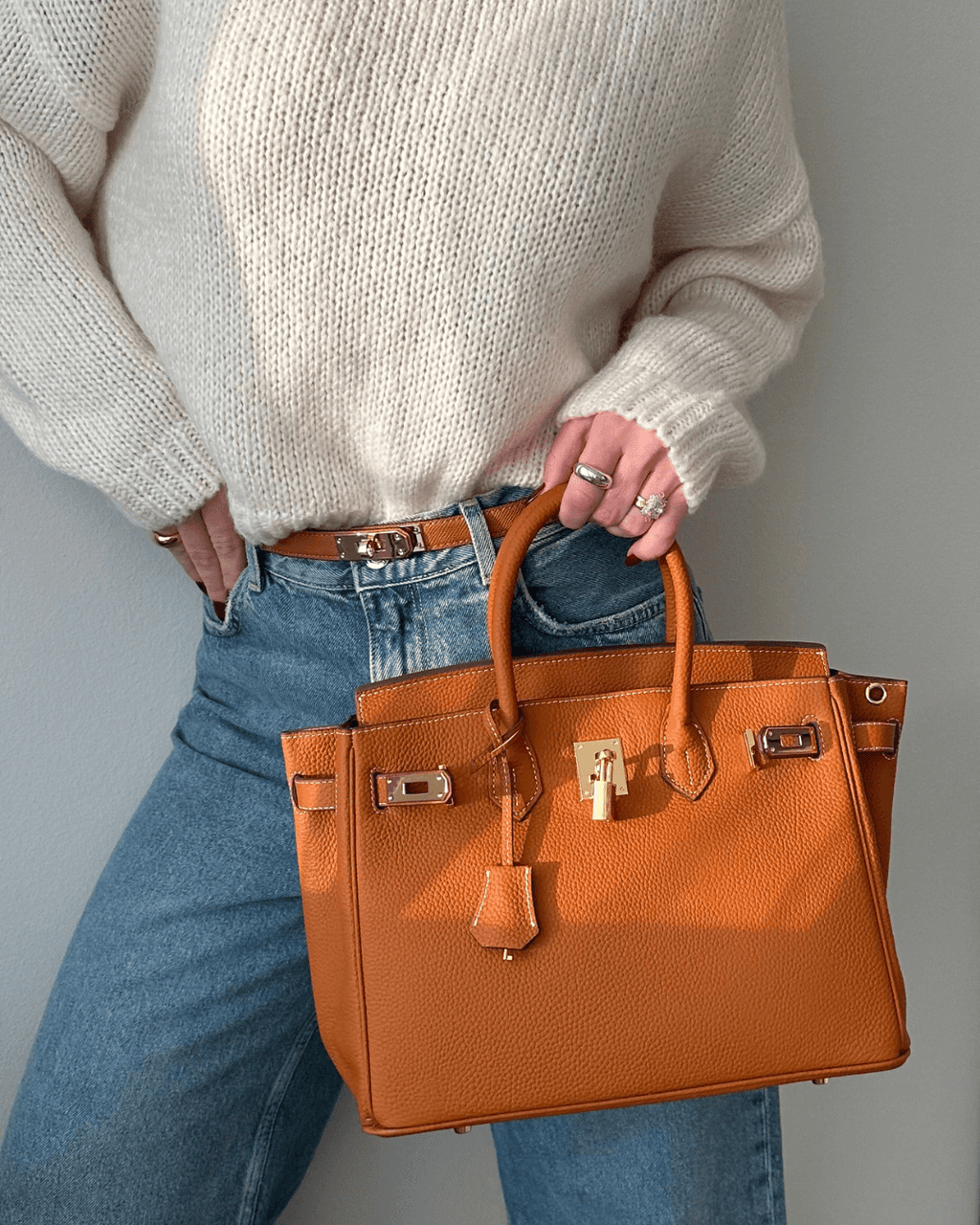 Femme portant un sac à main en cuir orange, style Birkin, avec un pull blanc et un jean. Mode élégante, accessoires de luxe, tendance automne.