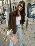 Femme souriante en veste marron, t-shirt blanc, jeans, portant lunettes de soleil, sac blanc et sac rose, rue urbaine avec vélos et feuilles d'automne.