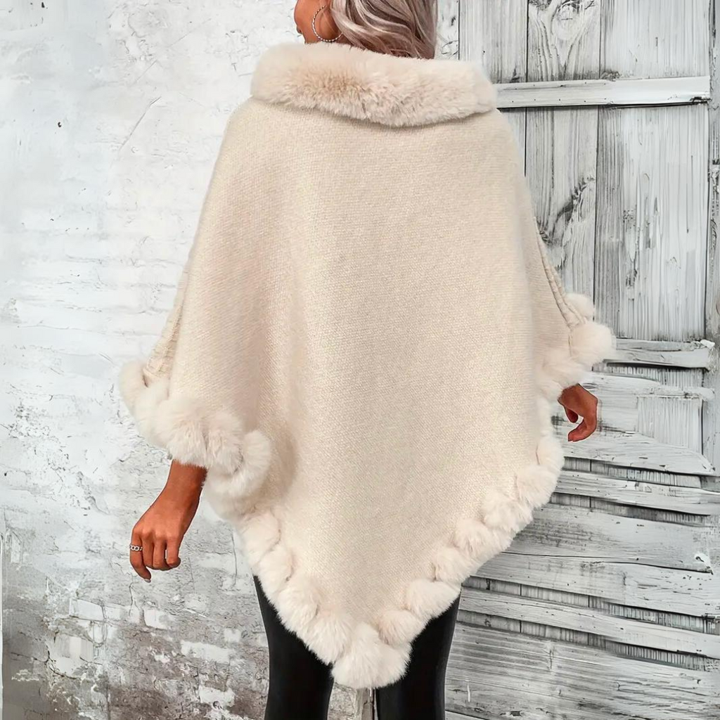 Femme portant un poncho beige en laine avec bordure en fausse fourrure, devant une porte en bois blanc. Mode hiver élégante et chic.