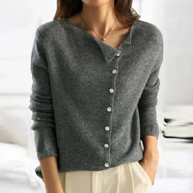 Femme portant un pull gris en laine avec boutons, manches longues, col asymétrique, style décontracté, sur fond intérieur lumineux.