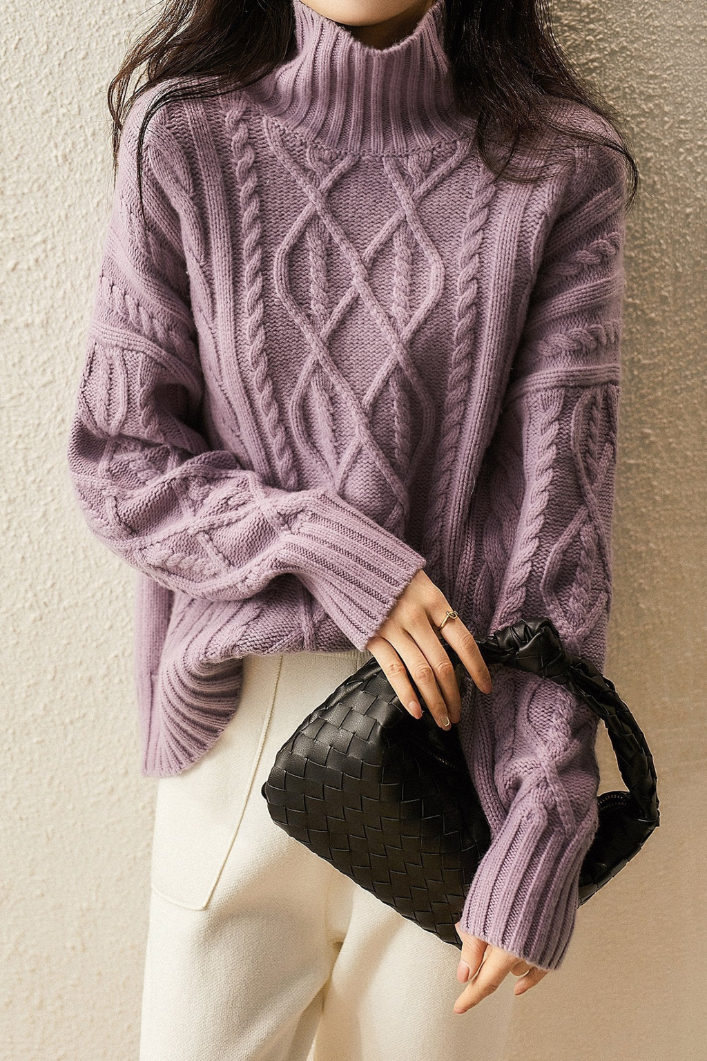 Femme portant un pull en laine violet à col roulé avec motifs torsadés, tenant un sac à main noir tressé. Mode automne-hiver élégante et tendance.