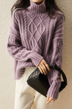 Femme portant un pull en laine violet à col roulé avec motifs torsadés, tenant un sac à main noir tressé. Mode automne-hiver élégante et tendance.