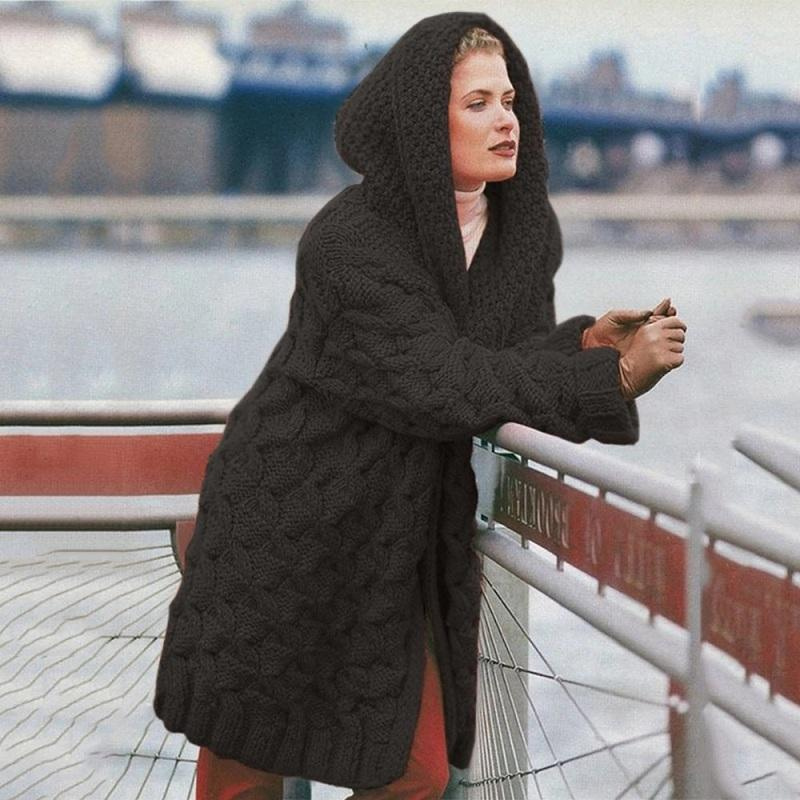 Femme portant un manteau en laine à capuche, tricot torsadé, debout près d'une balustrade avec vue sur un pont et une rivière en arrière-plan.
