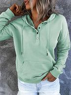 Femme portant un sweat à capuche vert menthe avec boutons, manches longues et poche kangourou, sur fond gris texturé. Mode décontractée tendance.