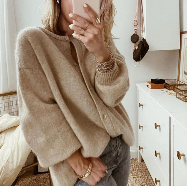 Femme prenant un selfie en portant un cardigan beige en laine, accessoires dorés, dans une chambre moderne avec commode blanche et décoration minimaliste.