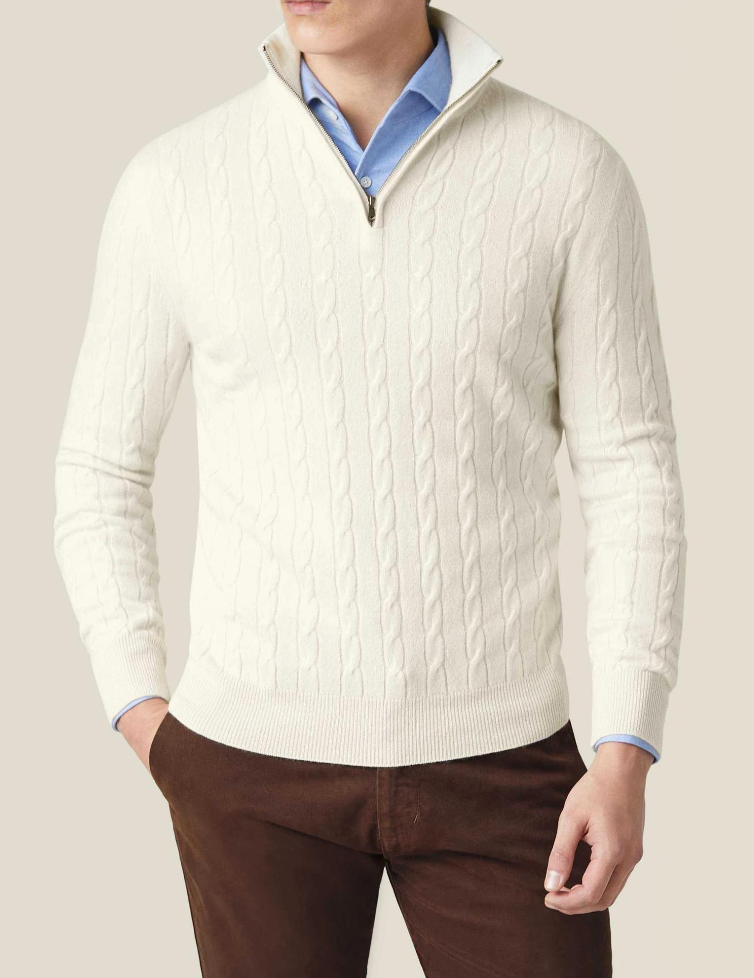 Homme portant un pull torsadé blanc à col zippé, chemise bleue et pantalon marron. Mode masculine élégante, tenue automne-hiver.
