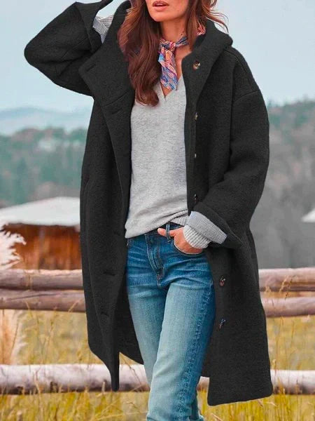 Femme portant un manteau noir oversize, pull gris, jeans bleu, foulard coloré, posant devant un paysage champêtre. Mode automne-hiver tendance.