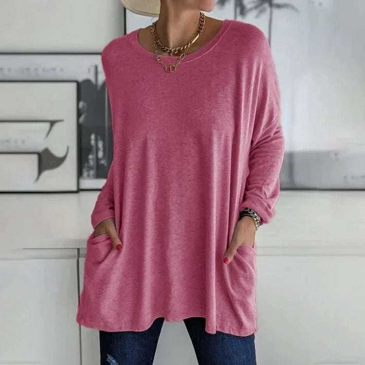 Femme portant un pull rose ample avec poches, collier doré, chapeau beige, devant un mur décoratif. Mode décontractée, tendance automne.