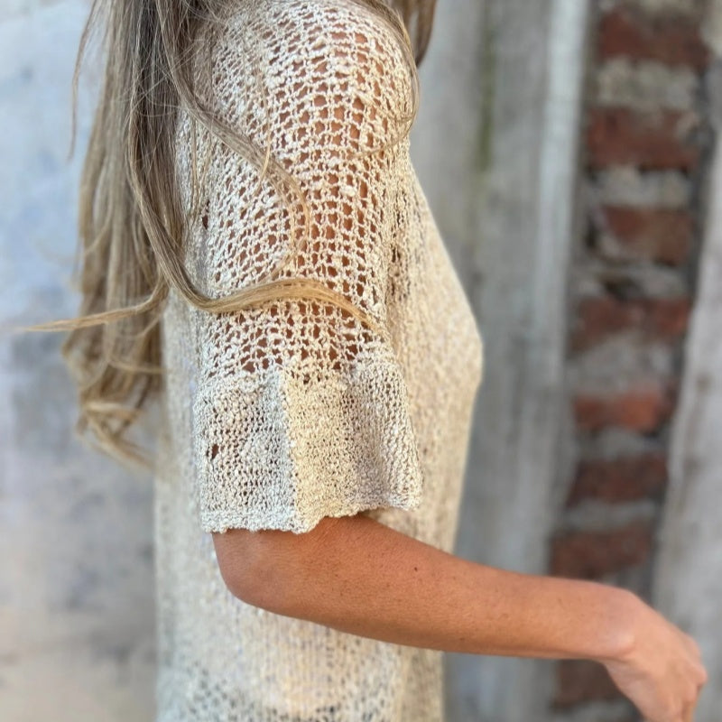 Femme portant un pull en maille beige, manches courtes, devant un mur en briques. Mode féminine, style décontracté, tendance tricot.