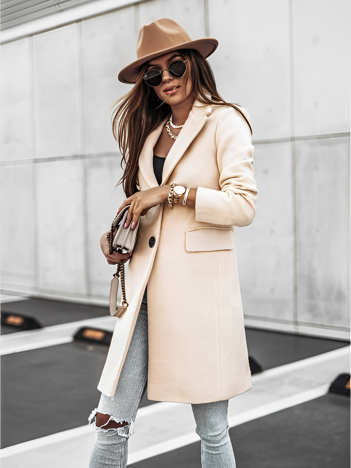 Femme élégante en manteau beige, chapeau marron et lunettes de soleil, tenant un sac à main, posant dans un environnement urbain moderne.