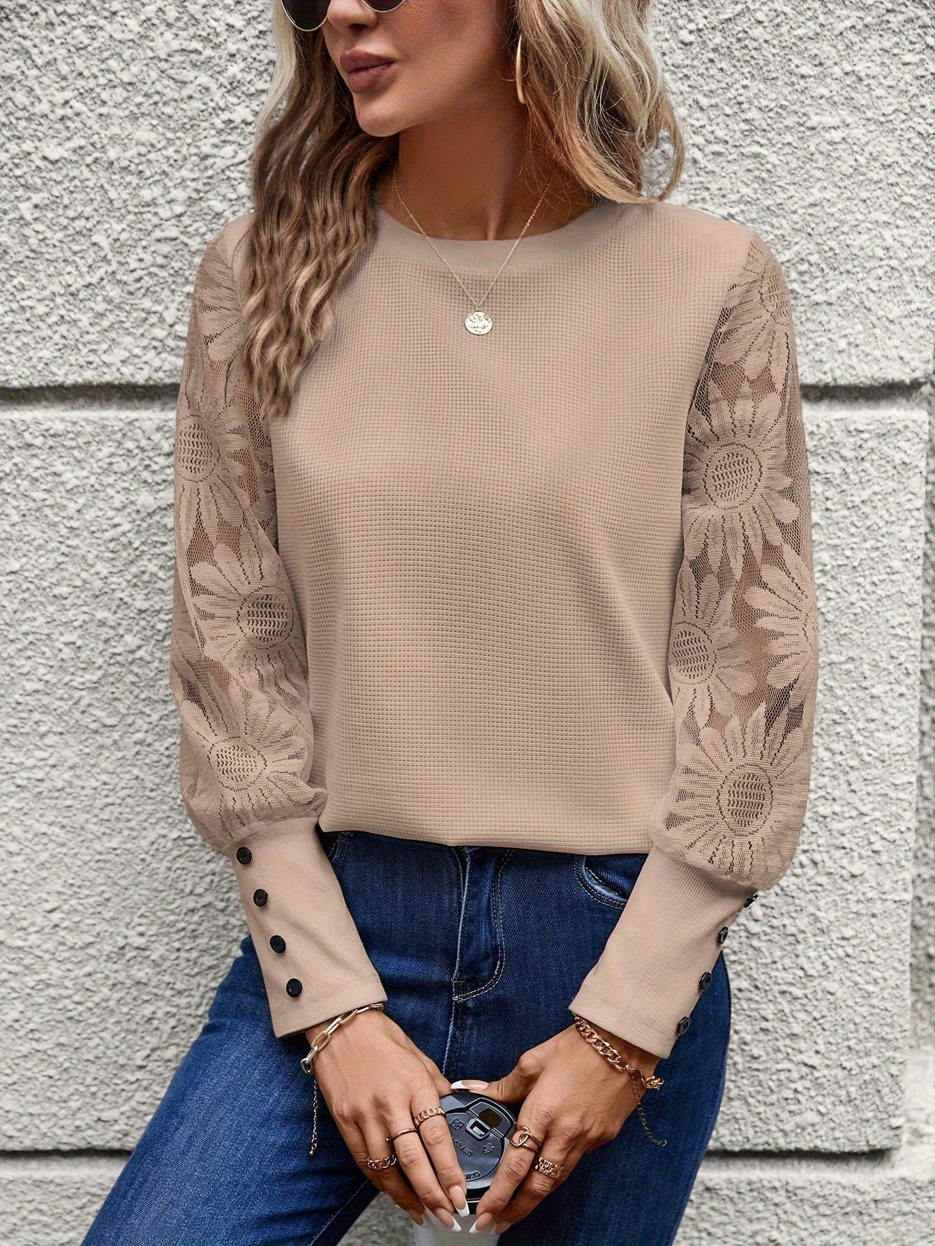 Femme portant un pull beige à manches longues en dentelle florale, boutons aux poignets, avec un jean bleu. Mode élégante et décontractée.
