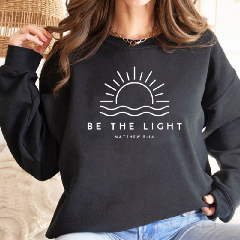 Femme portant un sweat noir avec un motif de soleil levant et l'inscription "Be the Light" et "Matthew 5:14". Mode décontractée, style inspirant.