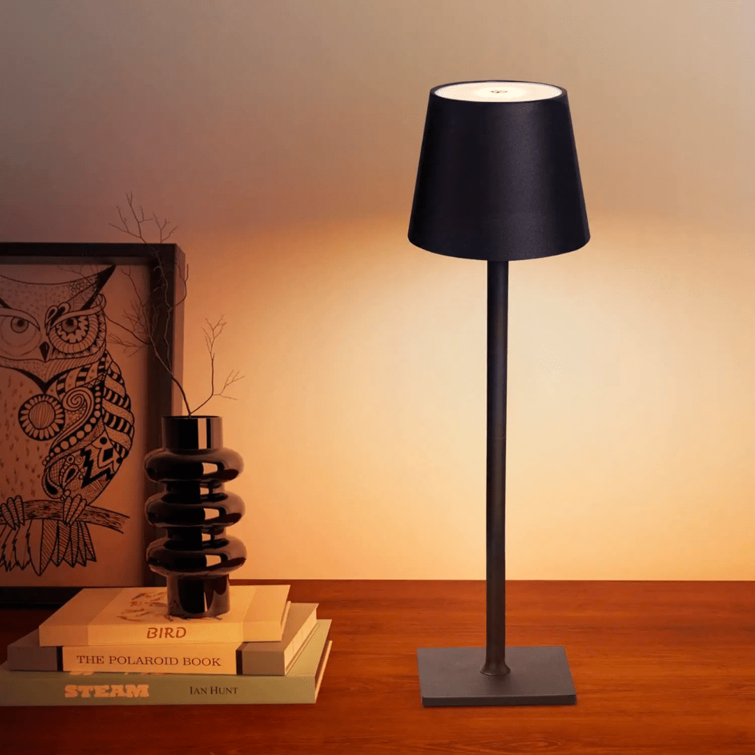 Lampe de table moderne noire sur bureau en bois, éclairant des livres empilés et un vase décoratif. Ambiance chaleureuse et élégante.