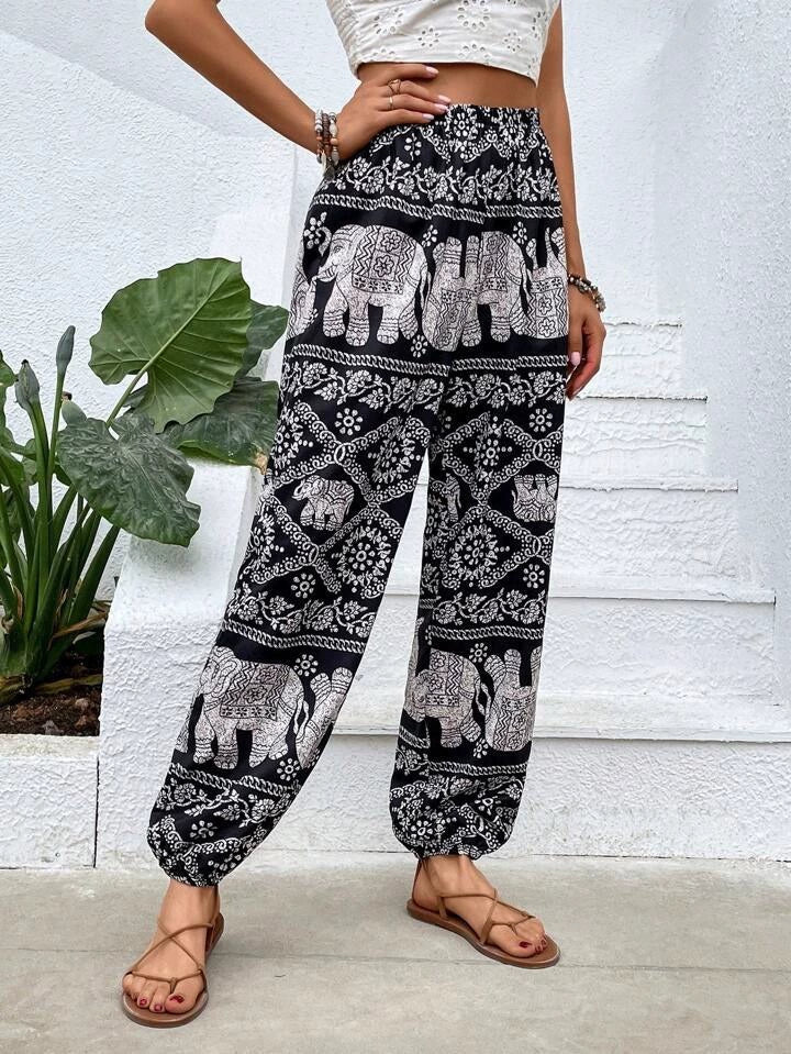 Pantalon ample noir et blanc avec motifs éléphants, femme debout, sandales marron, mur blanc en arrière-plan, plante verte à gauche.