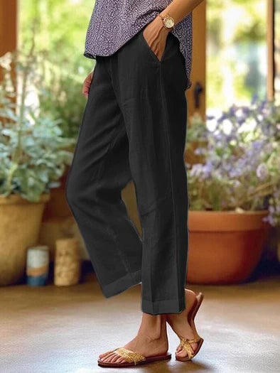 Pantalon noir en lin pour femme, style décontracté, poches latérales, porté avec des sandales dorées, ambiance estivale, plantes en arrière-plan.