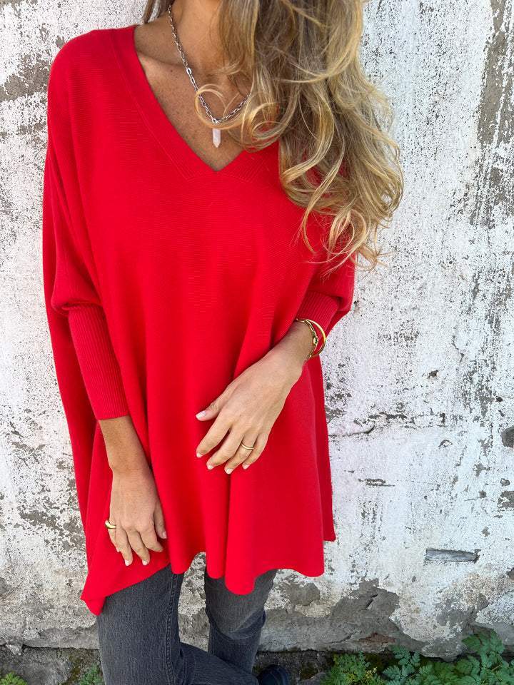 Femme portant un pull rouge ample à col en V, posant devant un mur texturé. Mode automne, style décontracté, cheveux ondulés.