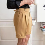 Homme portant un short beige élégant avec pinces, ceinture ajustable, chemise noire; style moderne, mode masculine, intérieur chic.