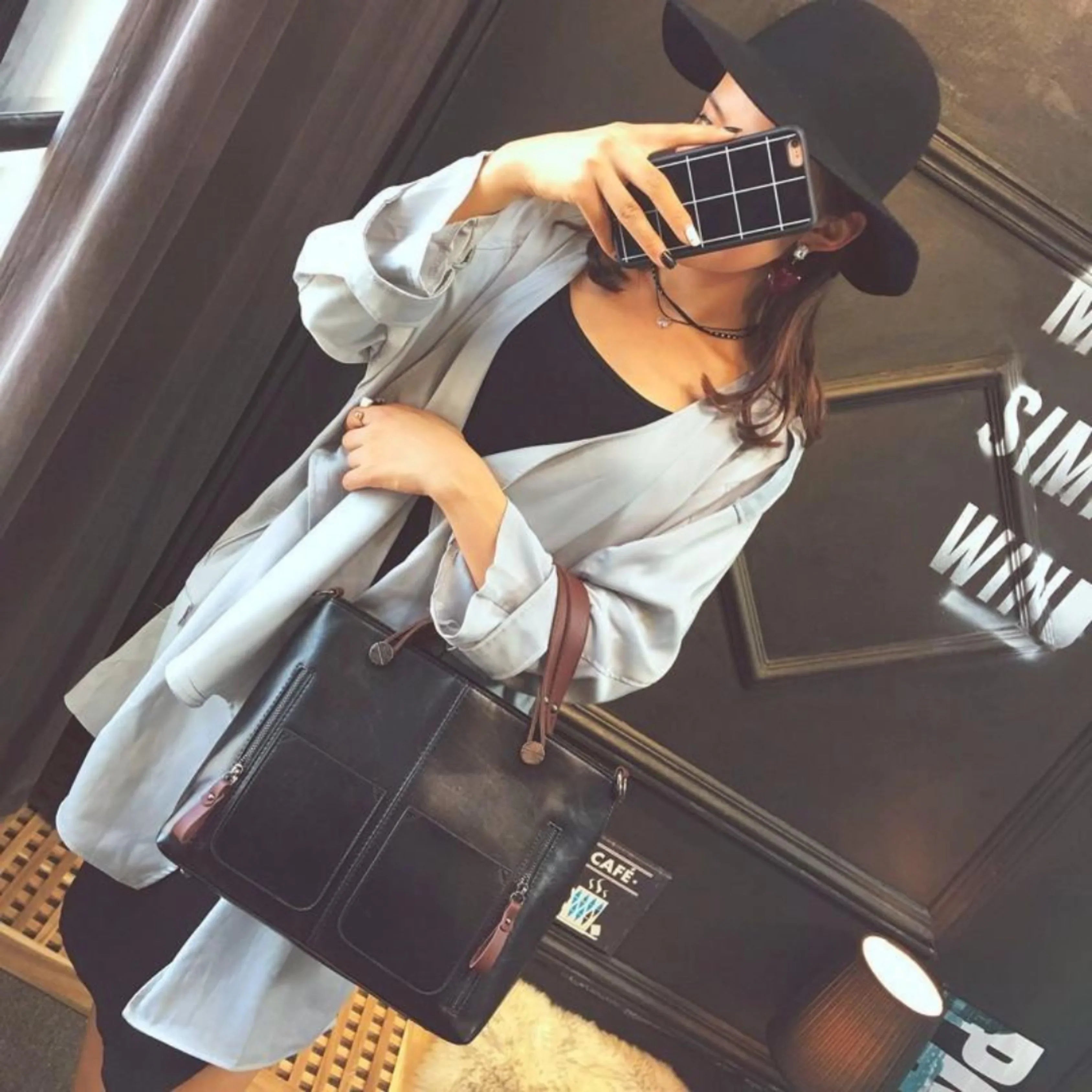 Femme élégante avec chapeau noir, manteau gris, sac à main en cuir noir, prenant un selfie avec un smartphone. Mode urbaine chic.