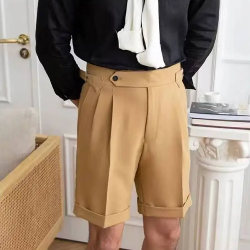 Homme portant un short beige élégant avec plis, chemise noire et écharpe blanche, debout dans un intérieur moderne. Mode masculine chic.