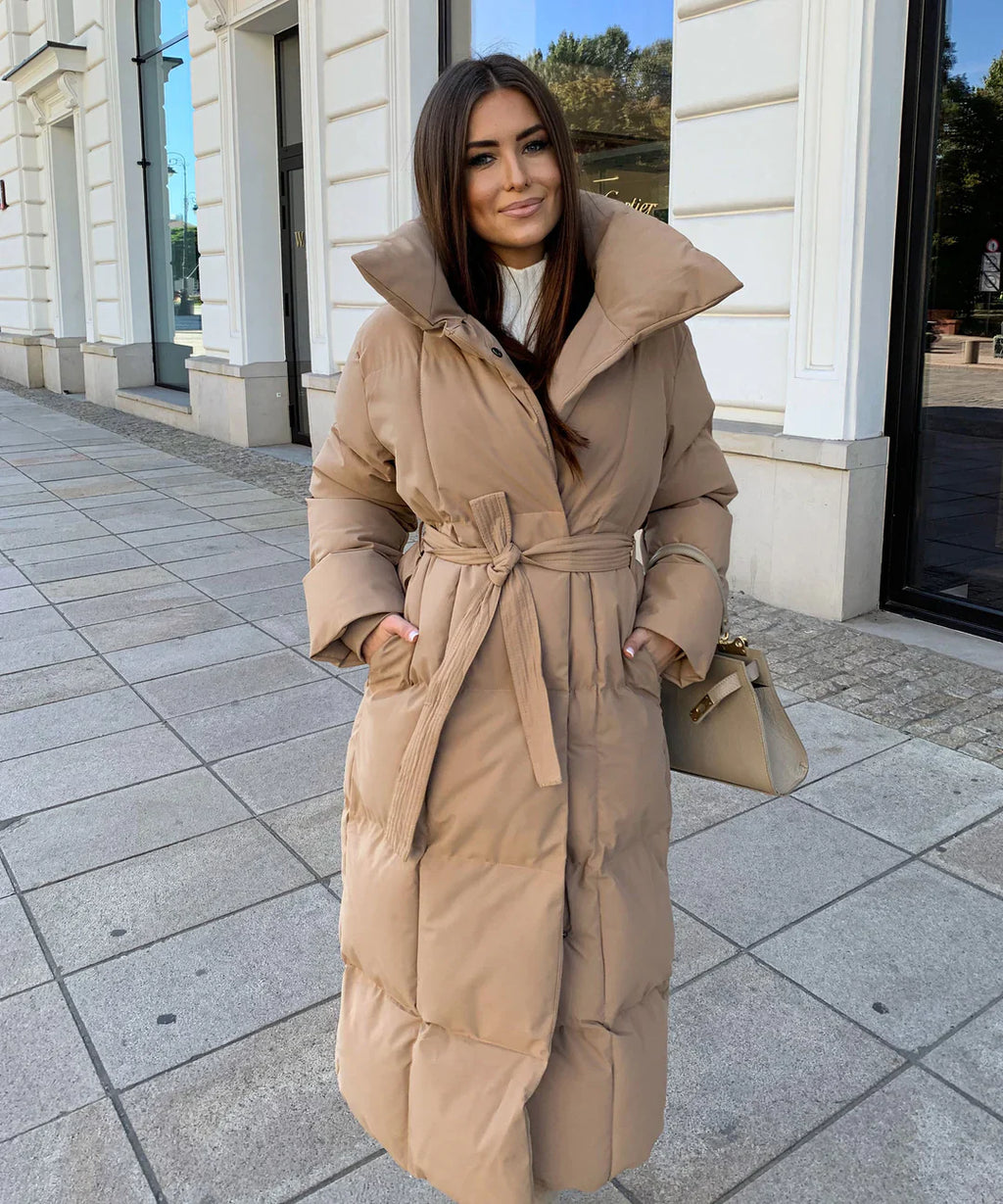 Femme portant un manteau long beige ceinturé, debout sur un trottoir urbain. Mode hiver élégante, tendance manteau matelassé.