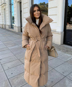 Femme portant un manteau long beige ceinturé, debout sur un trottoir urbain. Mode hiver élégante, tendance manteau matelassé.