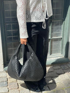 Femme tenant un sac à main noir tressé, portant un chemisier en dentelle blanche et un pantalon noir, debout sur des pavés devant une fenêtre.