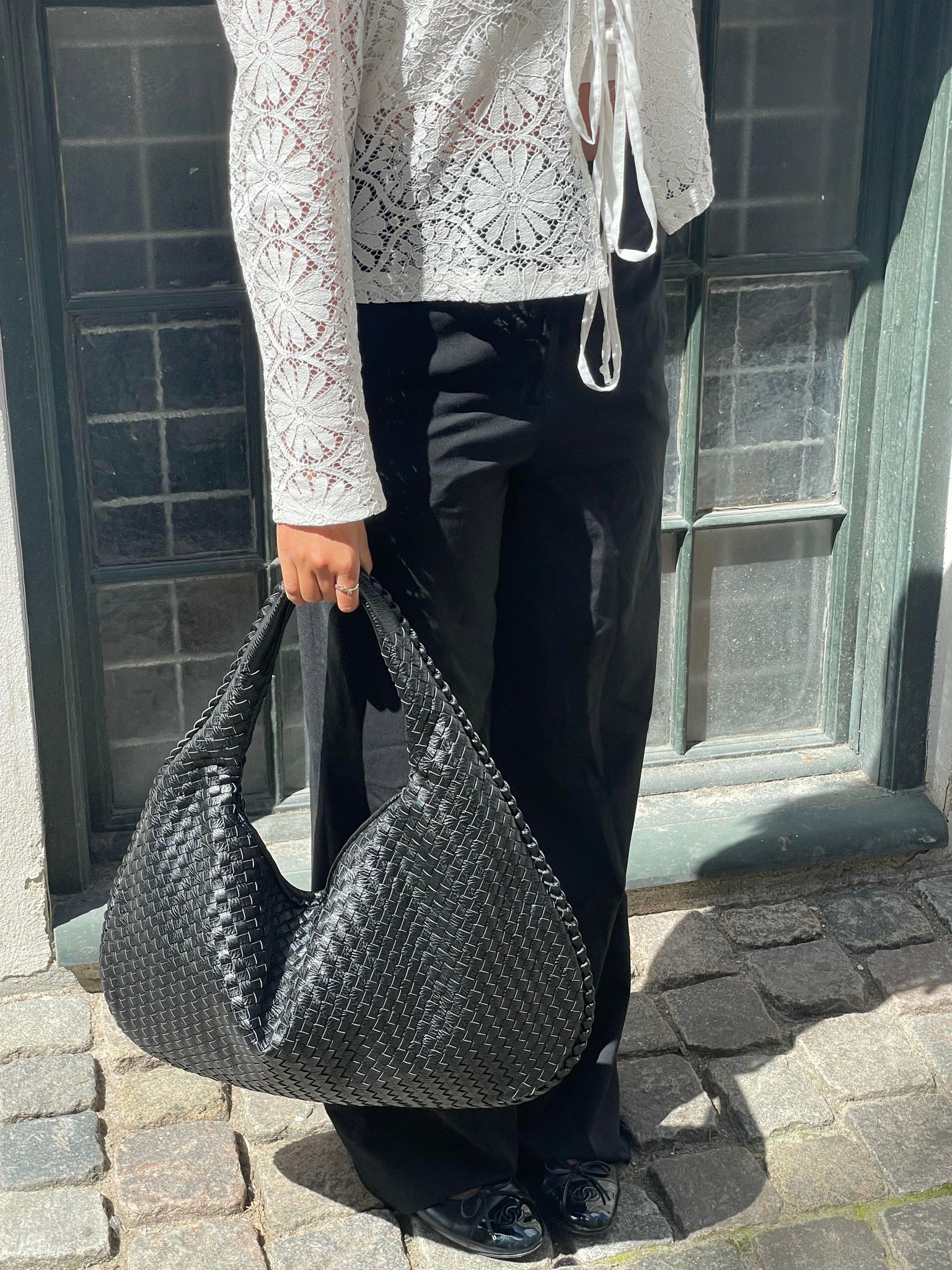 Femme tenant un sac à main noir tressé, portant un chemisier en dentelle blanche et un pantalon noir, debout sur des pavés devant une fenêtre.