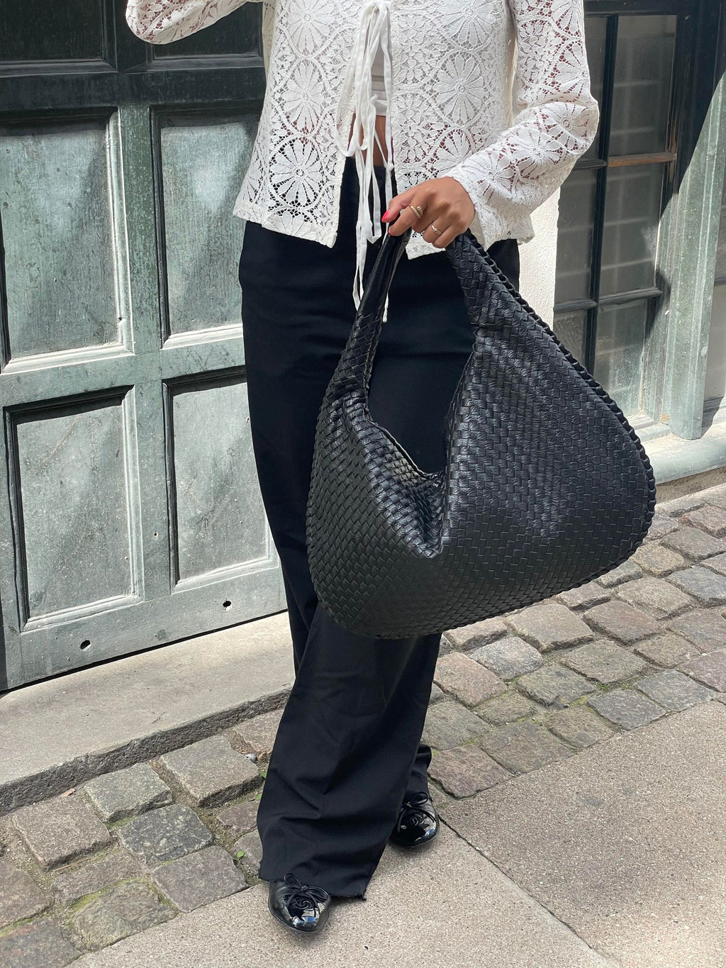 Femme portant un sac à main noir tressé, tenue élégante avec chemisier en dentelle blanche et pantalon noir, devant une porte en bois.