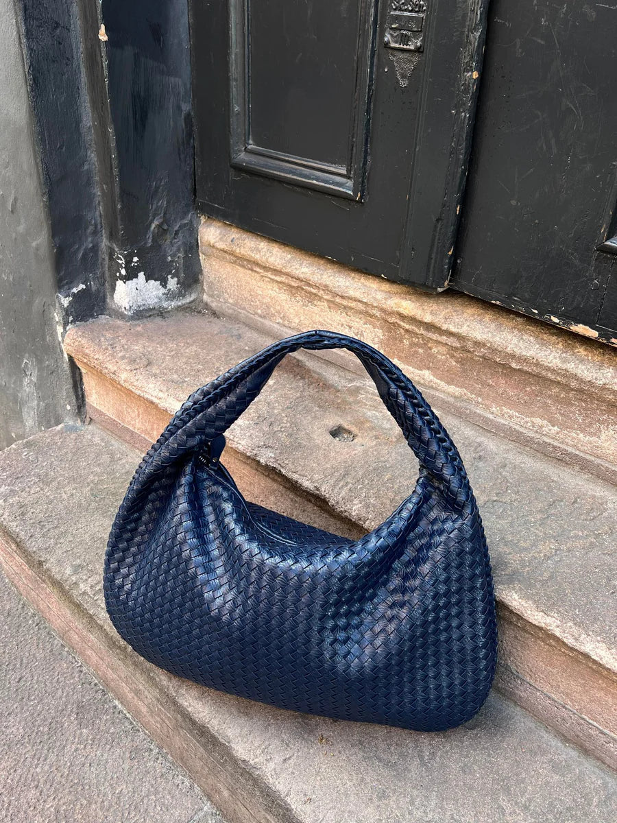 Sac à main bleu tressé posé sur des marches en pierre devant une porte noire. Accessoire mode élégant, tendance et chic pour femme.