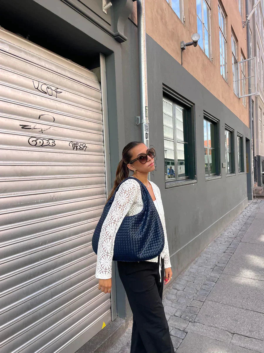 Femme portant un sac à main bleu, lunettes de soleil, chemisier blanc en dentelle, pantalon noir, posant devant un bâtiment urbain moderne.