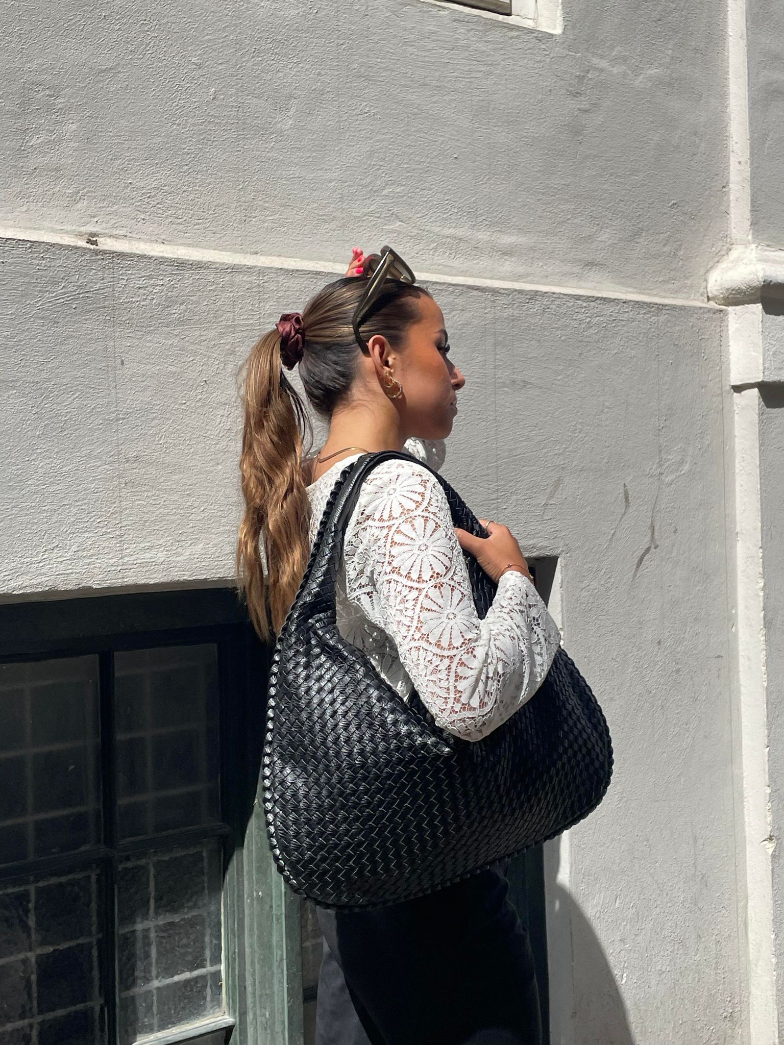 Femme portant un grand sac à main noir tressé, vêtue d'un chemisier blanc en dentelle, posant devant un mur blanc. Mode urbaine élégante.